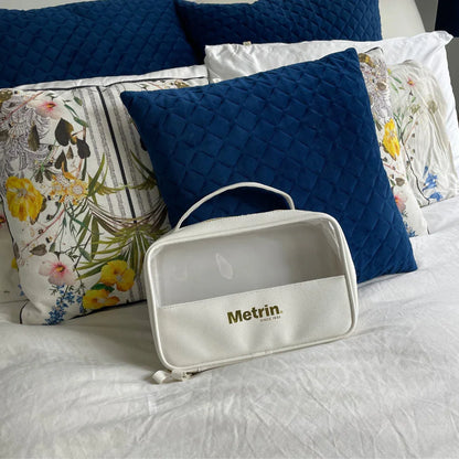 White toiletry bag with 'Metrin' branding on a bed with blue and floral-patterned pillows.